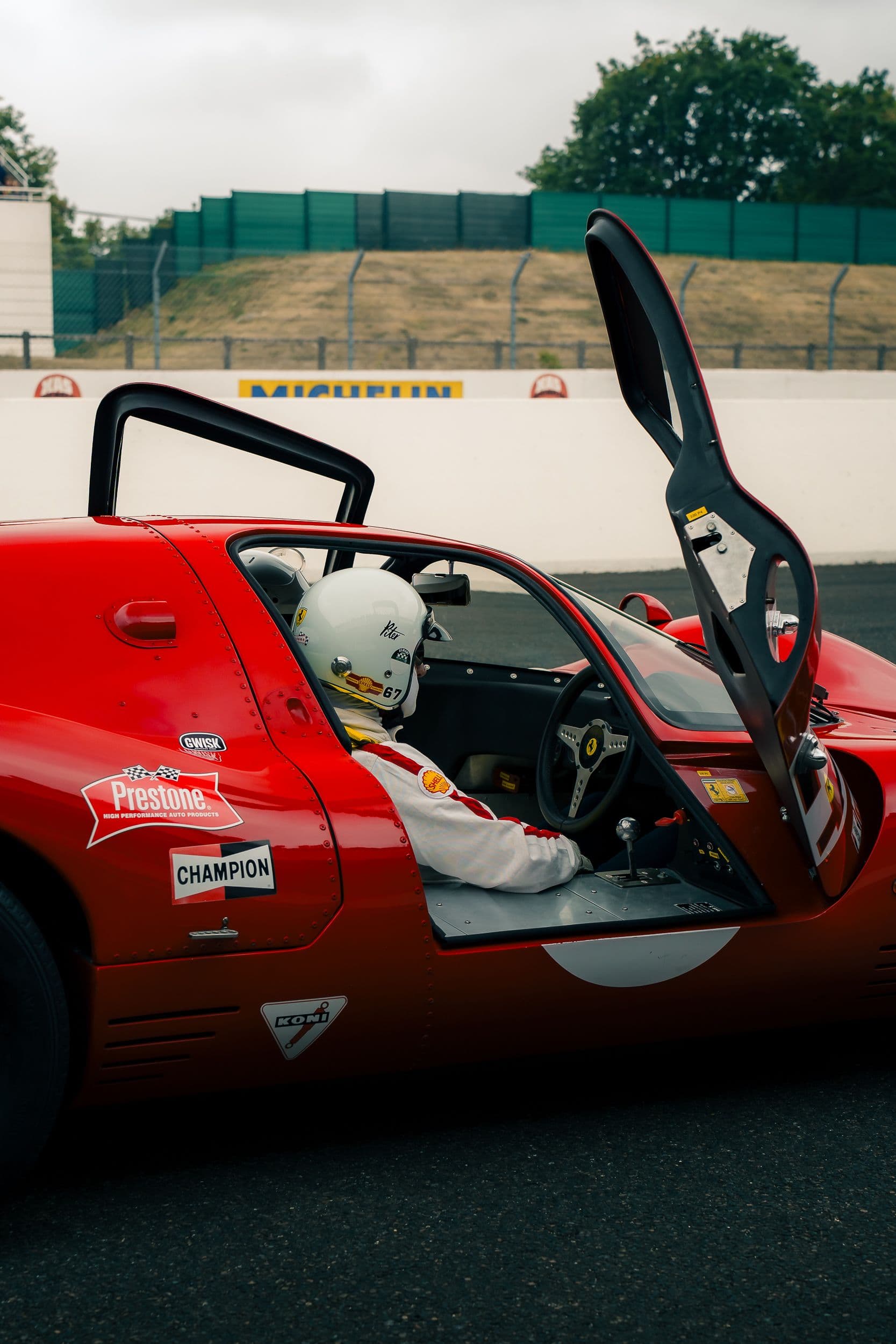 Pilote au casque blanc dans une Ferrari 330 P4 Replica rouge, porte relevée et volant visibles.