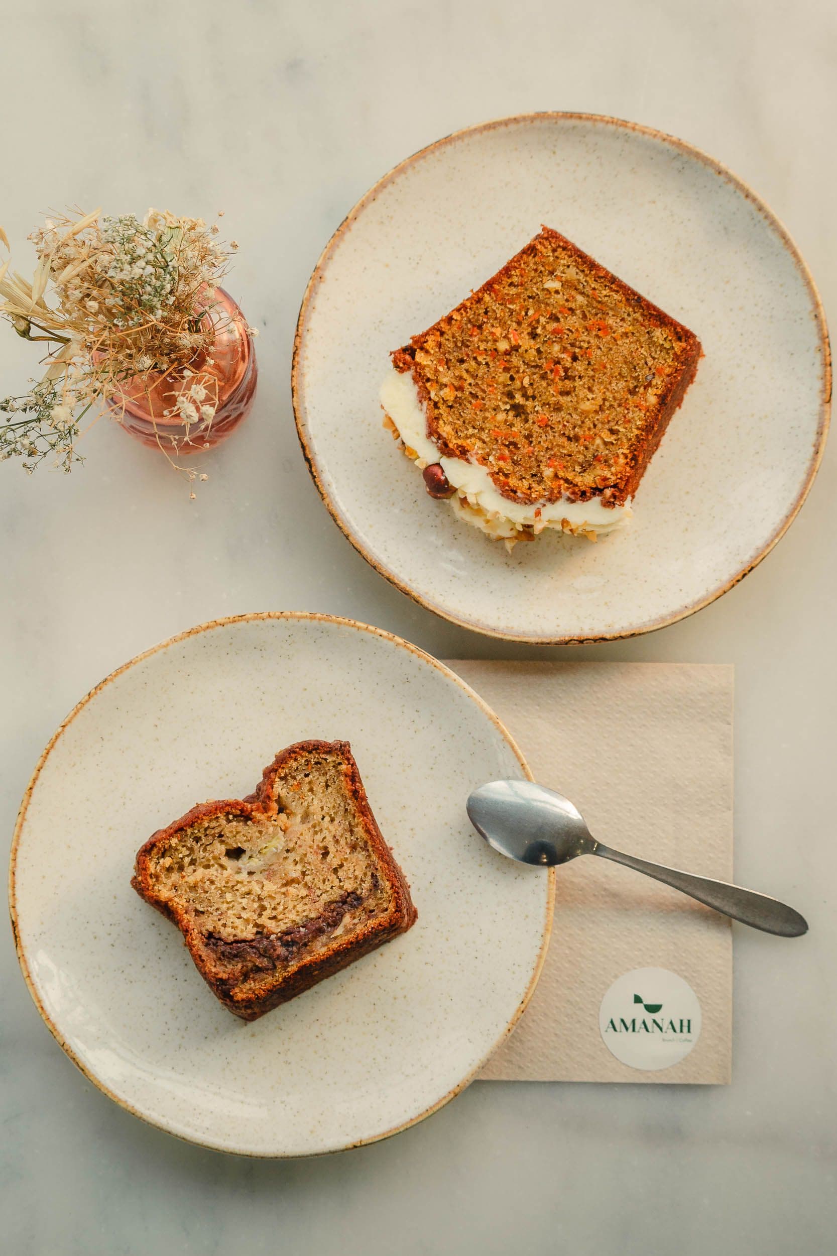 Tranche de carrot cake artisanal avec glaçage et fleurs séchées en décoration, photographie culinaire pour Amanah Coffee Shop