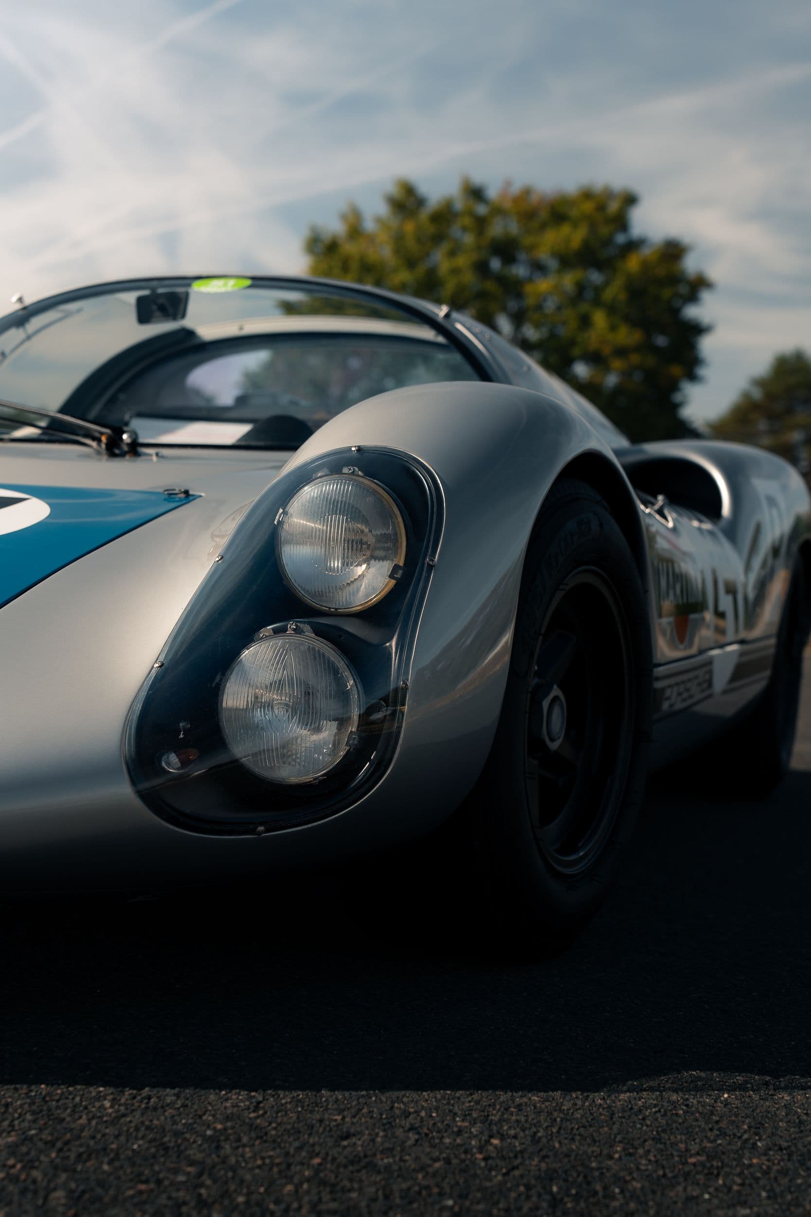 Gros plan sur les deux phares superposés sous une protection transparente d’une Porsche 910 argentée.