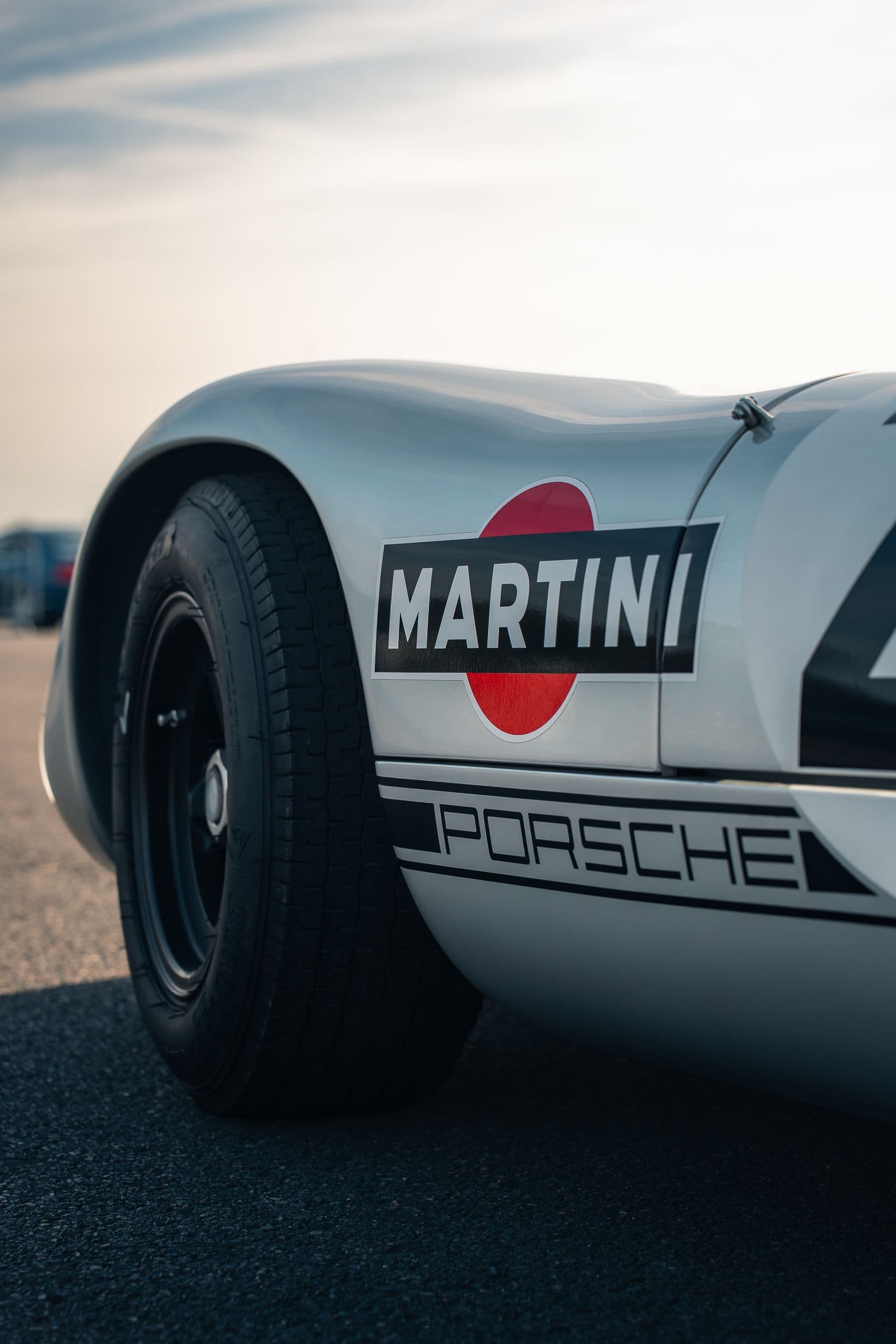 Gros plan sur le logo Martini et la roue avant noire d’une Porsche 910 argentée.