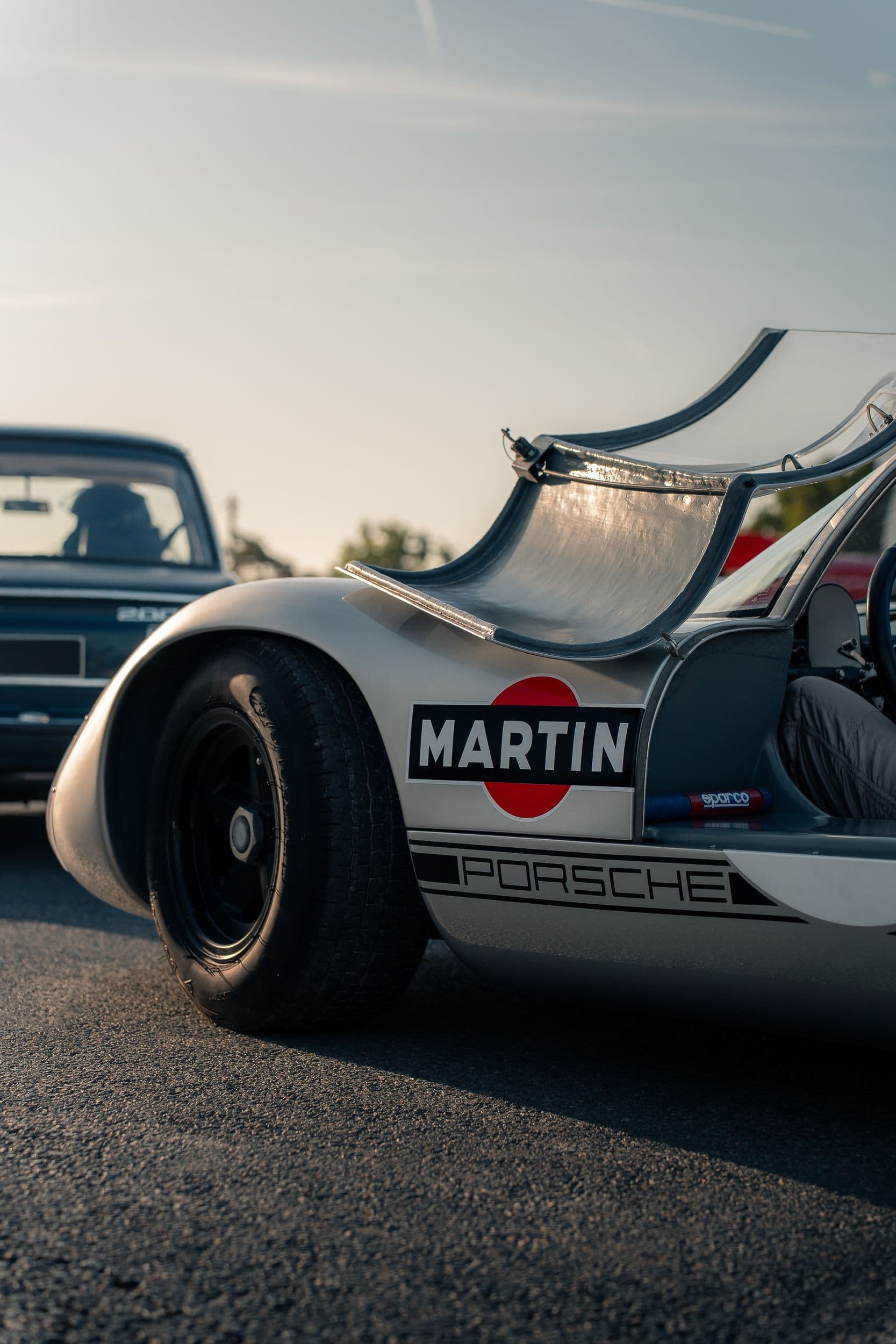 Porte relevée d’une Porsche 910 argentée, avec roue avant noire et logo Martini.