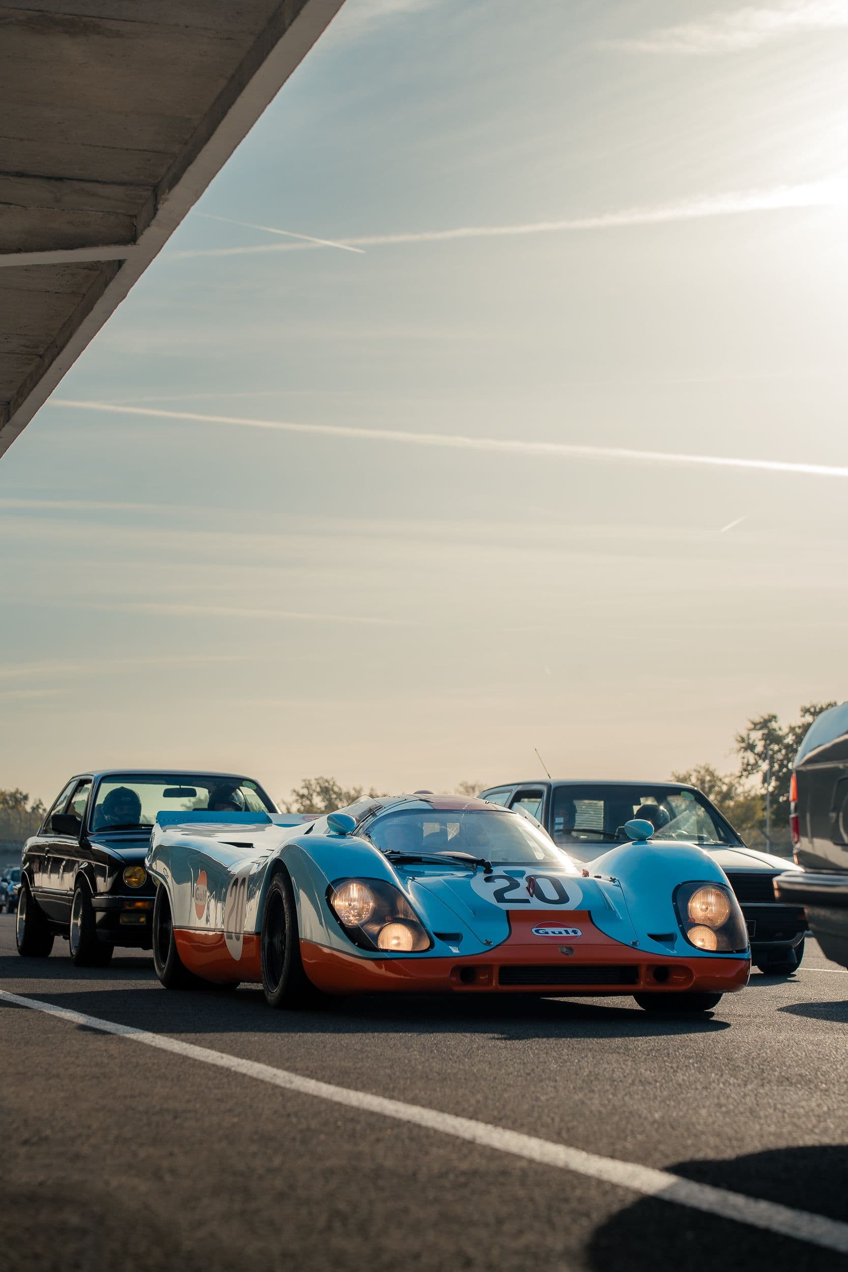 Porsche 917 Replica en livrée Gulf, phares allumés, entourée de voitures dans une lumière rasante.