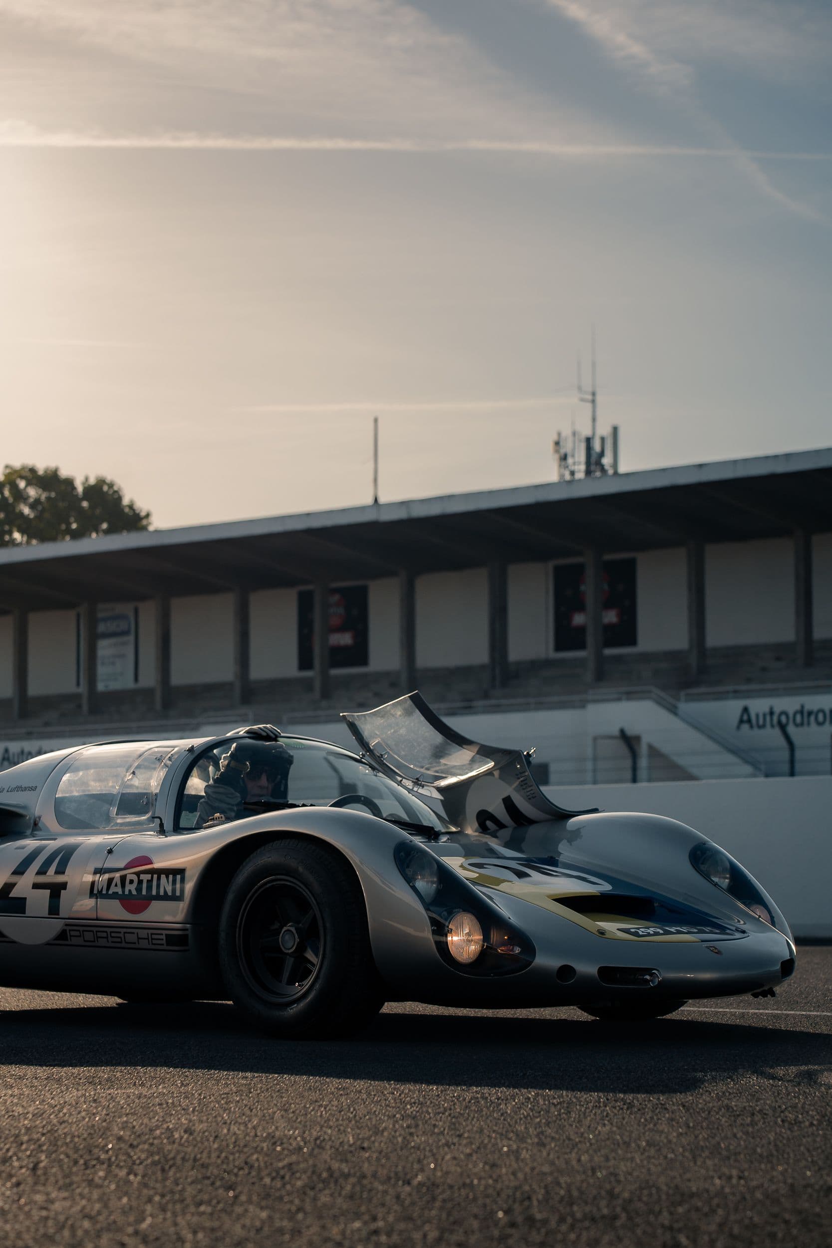 Porsche 910 argentée vue de trois quarts avant, porte ouverte et pilote casqué à bord.