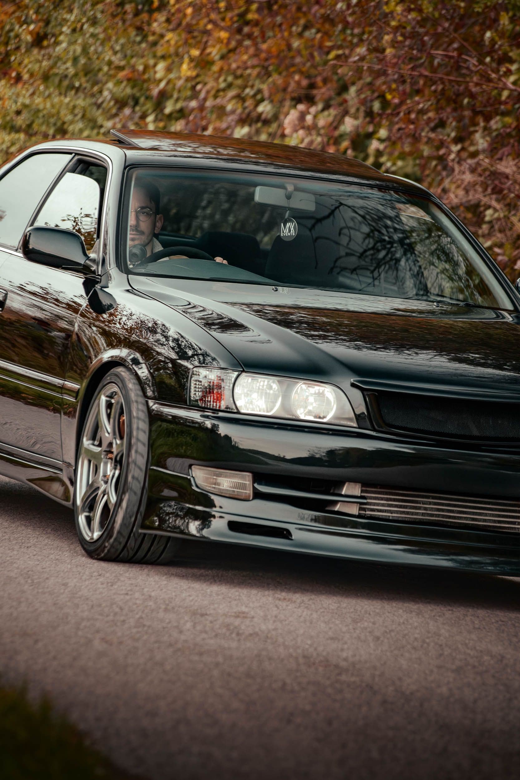 Toyota Chaser JZX100 trois-quarts avant en mouvement avec conducteur visible, berline JDM roulage automne, photographie dynamique