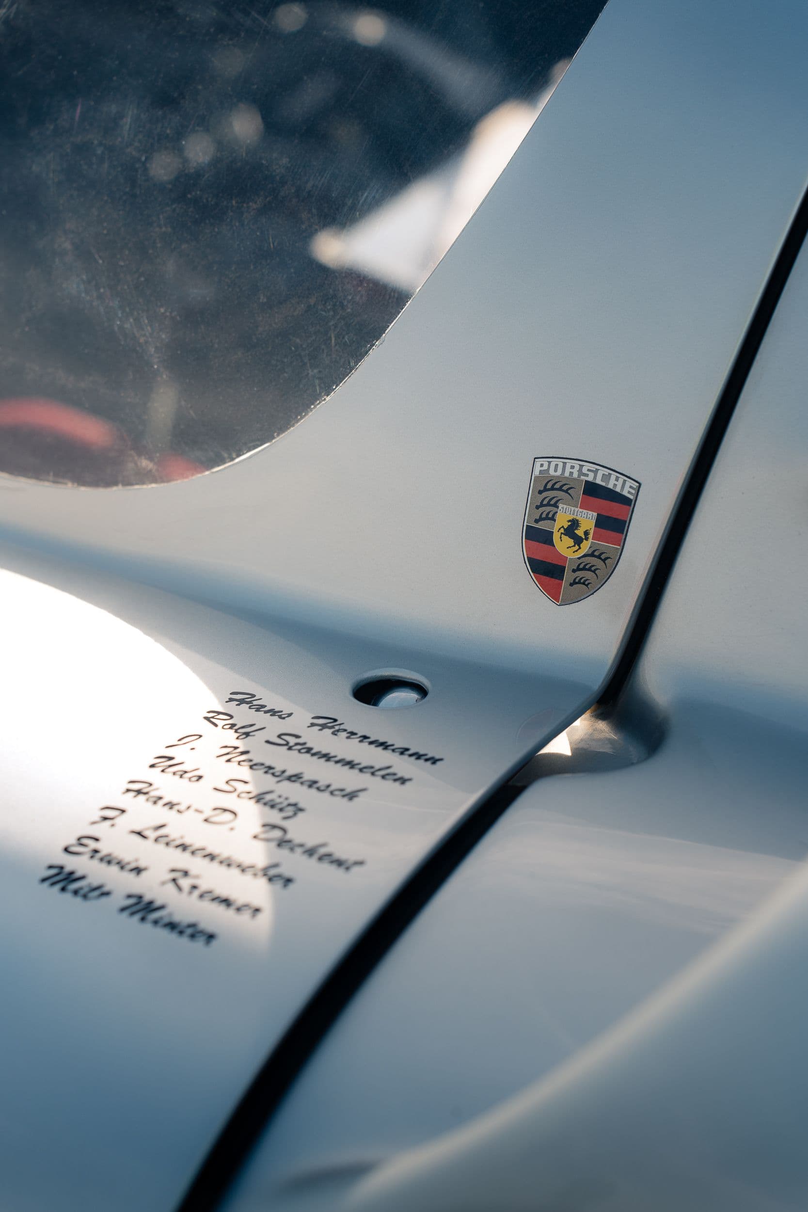 Écusson Porsche et noms de pilotes inscrits sur la carrosserie argentée près du vitrage.