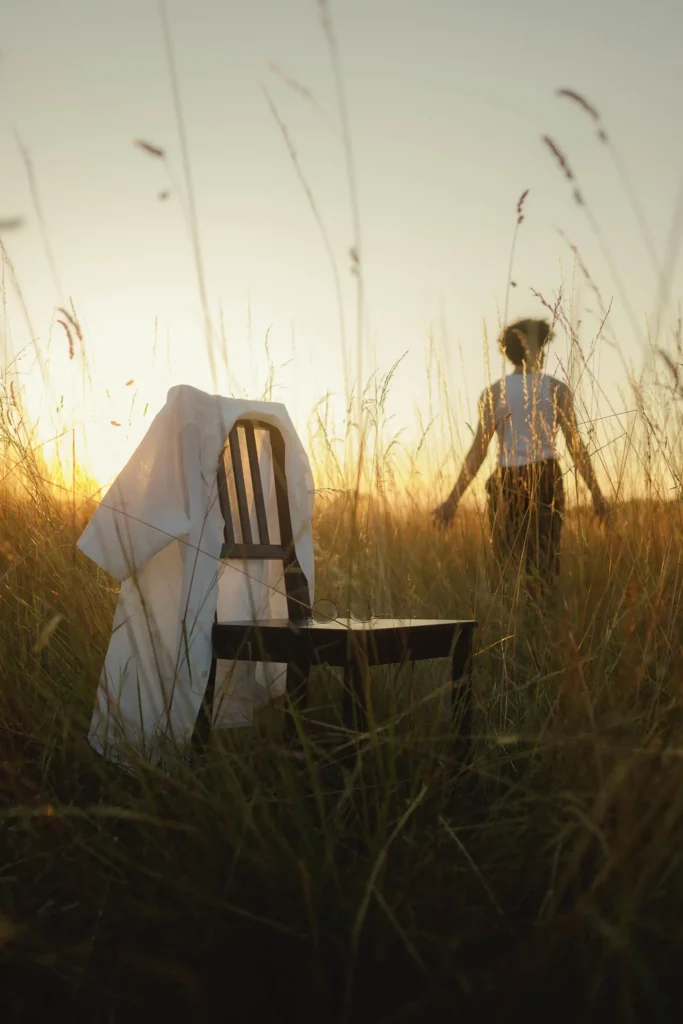 Une chaise et une chemise posées dans un champ, silhouette en arrière-plan au soleil couchant, atmosphère poétique.